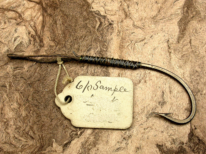 14a Unknown make hook, 6/0, forged, bronzed, turned up taper, snelled with bronze wire. This is a beautiful little hook. Almost certain to be of English make. Ca 1900’s. Original hanging tag reads, 6/0, Sample. Whatever else the tag said has been erased. Total length of hook and snell is 3 ¼”. 1900’s collection.