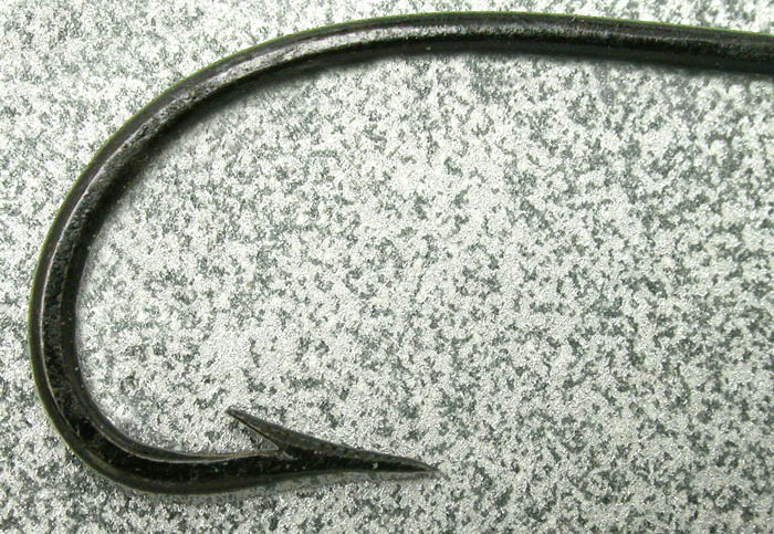Unidentified hook, about 1/0, forged, Marked, knobbed, flared, japanned. This is a very interesting little hook. It exhibits no less than three methods of snell retention. It is flared right behind the knob, has the knob as well and is marked. The markings are only on top of the shank which is rare. The marks are hand filed. How can you tell? Ground marks will be round at the bottom of each mark. Embossed marks will tend to be rounded top and bottom and file cut marks will be sharp top and bottom. Also, as in this hook, the area behind the flared area, is not flared which embossing will do.  The hook point is guttered and is a very nice Dublin point. Gift from Martin Bach